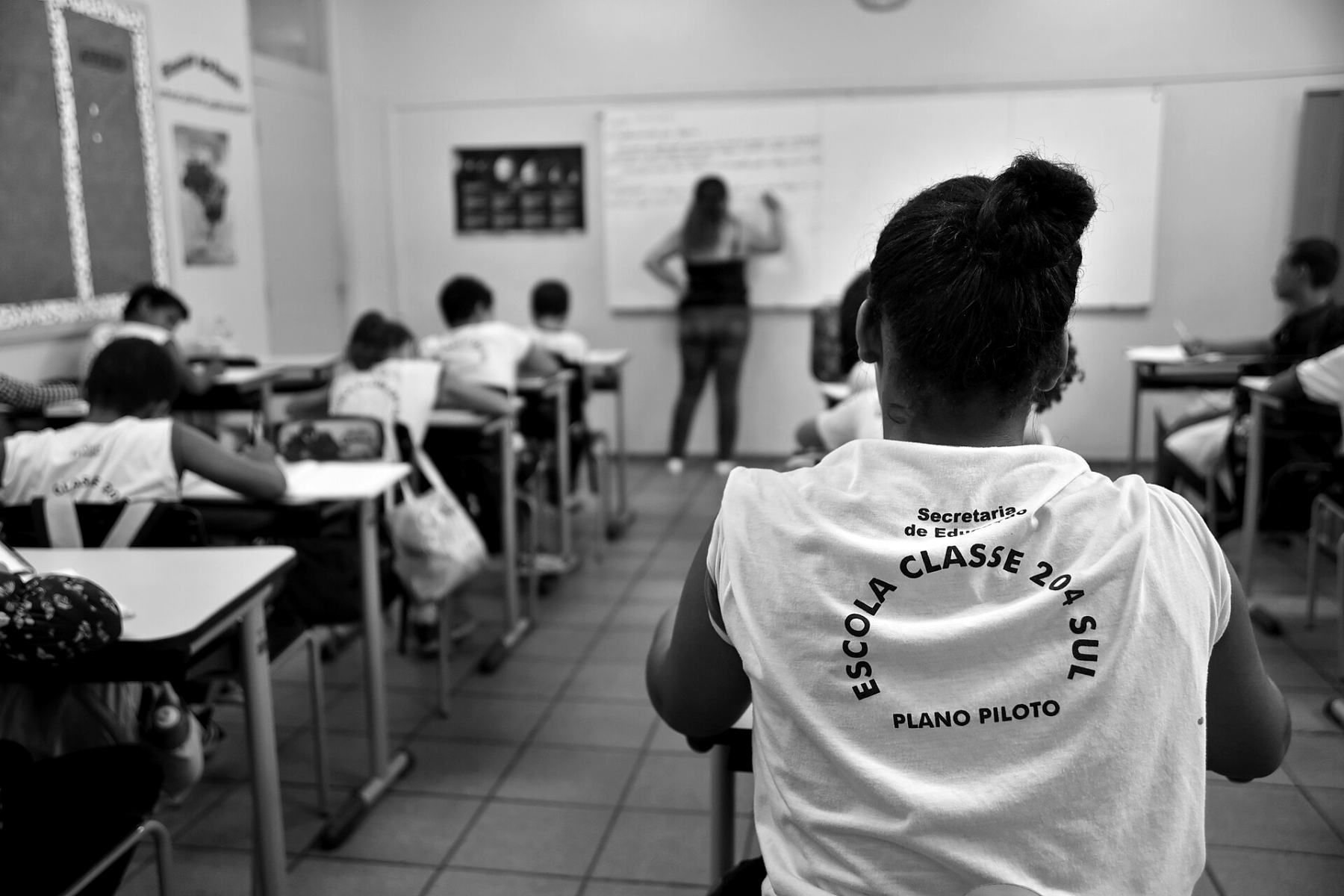 Sala de aula de escola pública, estudantes vistos de costas, em preto e branco